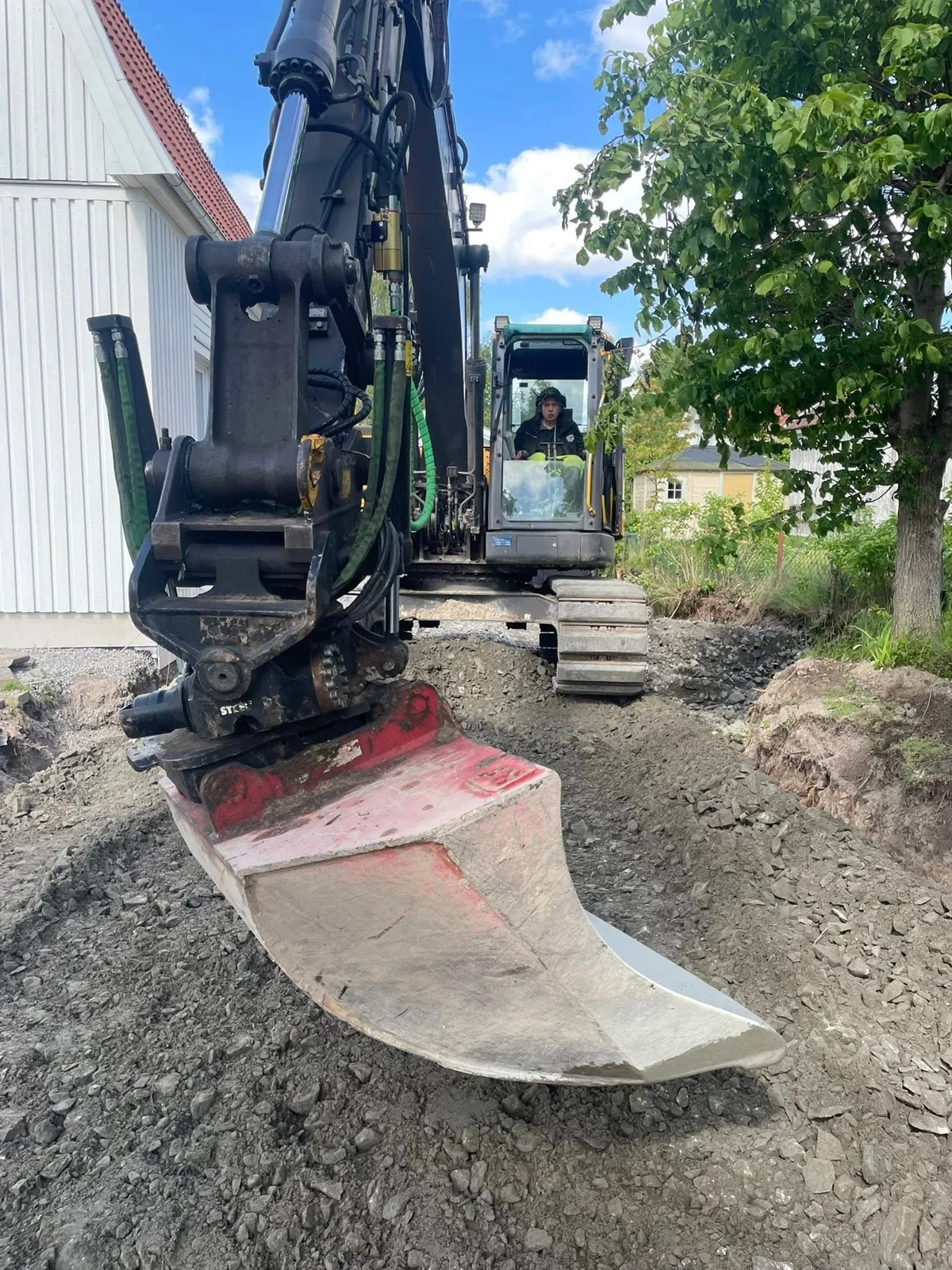 Grävmaskin med planeringsskopa på en villatomt, föraren syns i hytten